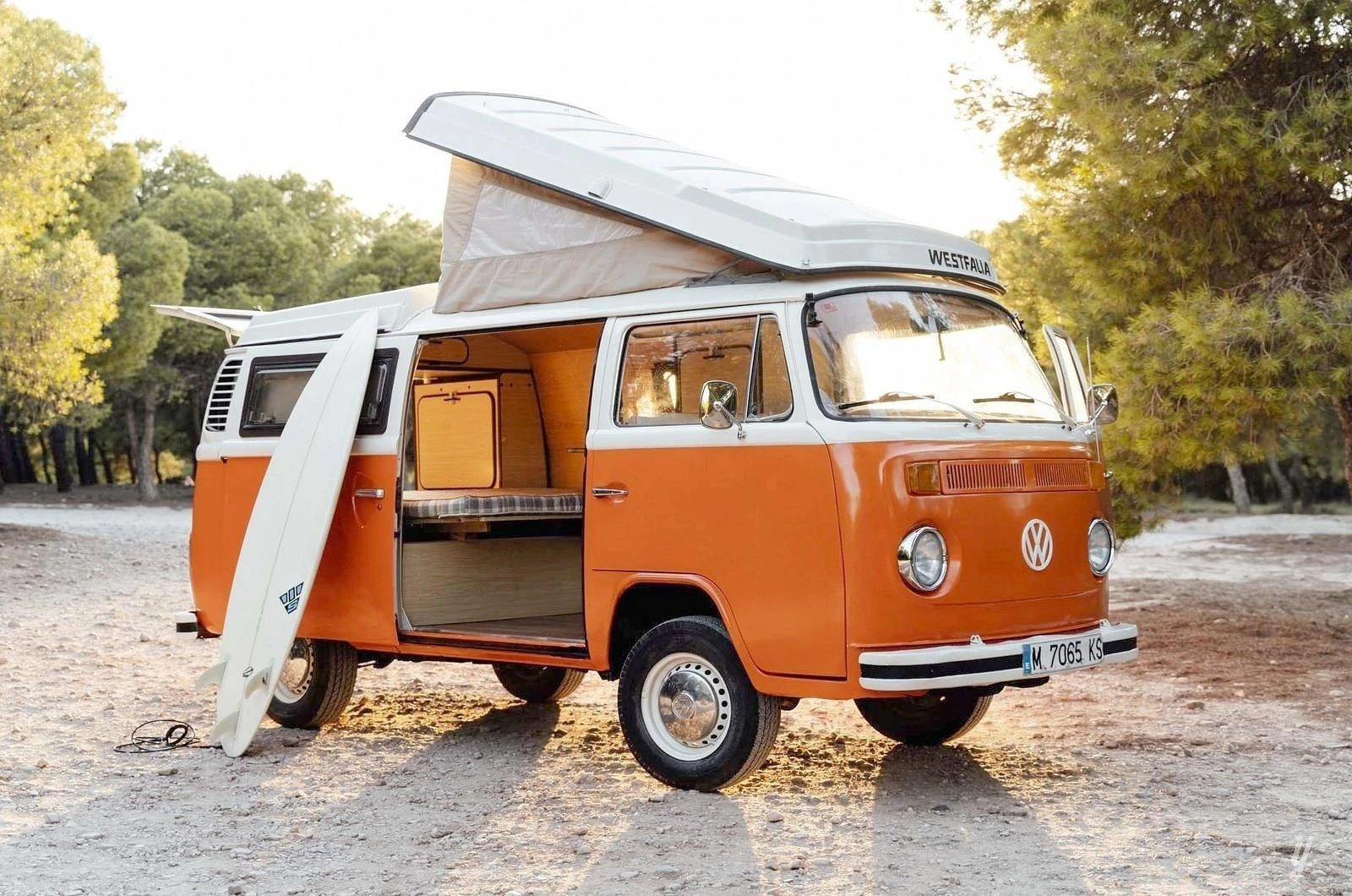 Combi Volkswagen T2 Westfalia orange et blanc, toit relevé et porte latérale ouverte, une planche de surf posée contre le flanc, sur une piste en forêt au coucher du soleil